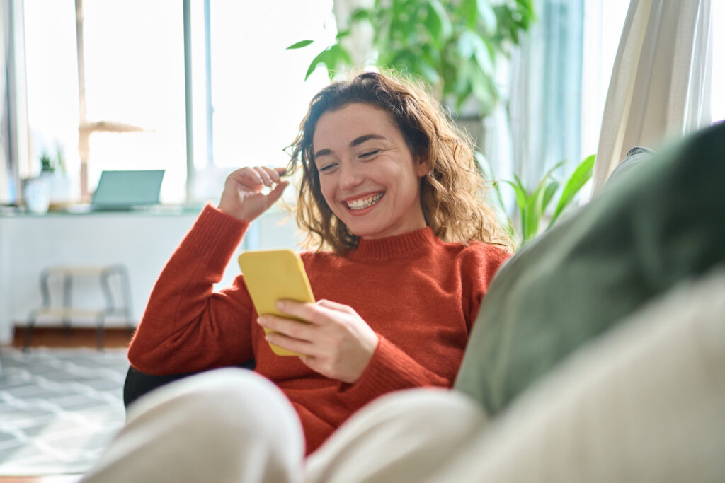 Young woman smiling while looking at her smartphone on a living room couch.